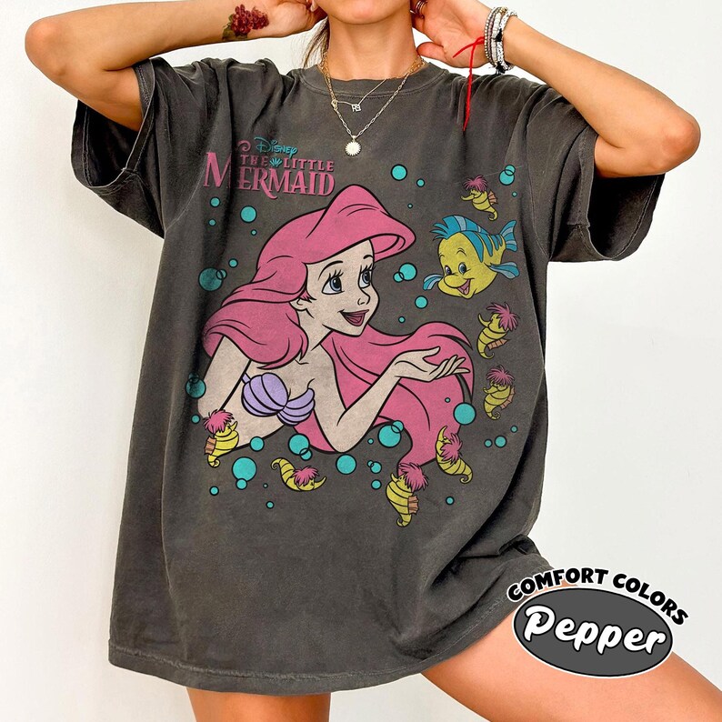 Puede incluir: Una camiseta gris carb&oacute;n de manga corta con un gr&aacute;fico de Ariel de La Sirenita con largo cabello rosa, un top de conchas morado y un pez amarillo. El dise&ntilde;o incluye burbujas verde azulado y peque&ntilde;as criaturas marinas amarillas sobre fondo oscuro.
