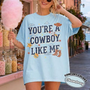May include: A light blue short-sleeve t-shirt featuring the dark blue text "YOU'RE A COWBOY LIKE ME" surrounded by graphic illustrations of a cowboy hat, a rocket, a sheriff star, a ball, and cowboy boots, with small clouds and stars.