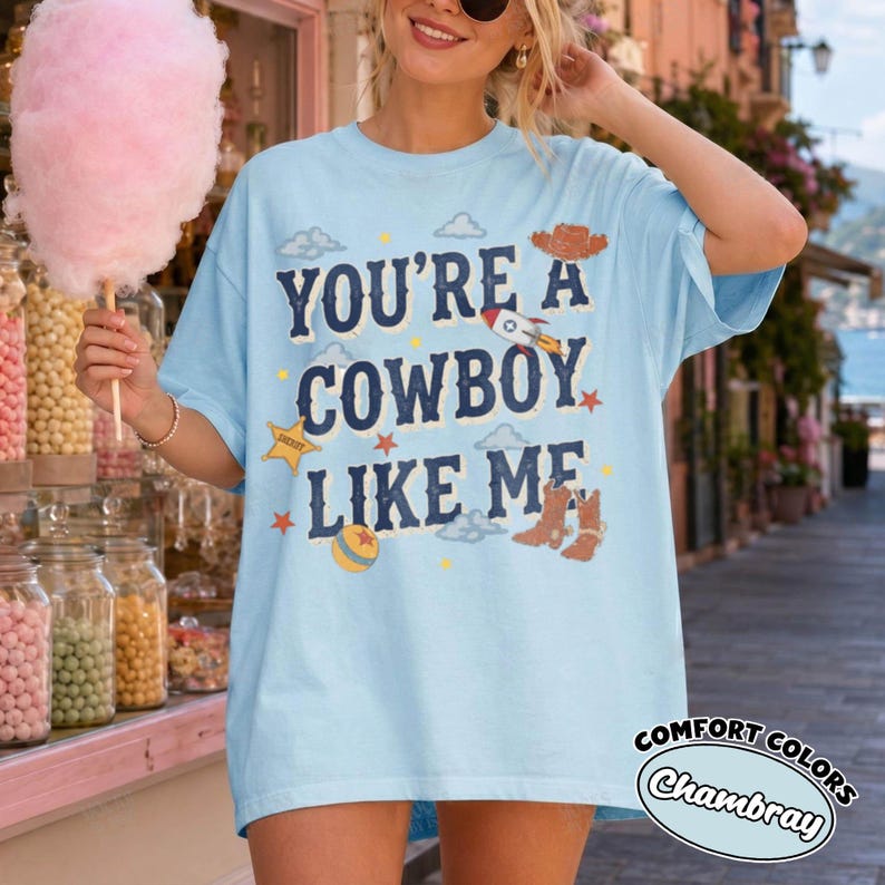 May include: A light blue short-sleeve t-shirt featuring the dark blue text "YOU'RE A COWBOY LIKE ME" surrounded by graphic illustrations of a cowboy hat, a rocket, a sheriff star, a ball, and cowboy boots, with small clouds and stars.