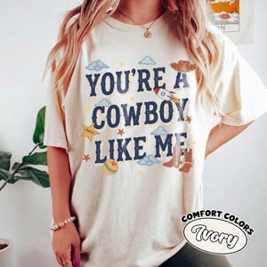 May include: An ivory short-sleeve t-shirt featuring the dark blue text "YOU'RE A COWBOY LIKE ME" surrounded by graphic illustrations of a rocket, a sheriff star, a ball, clouds, a cowboy hat, and a pair of brown cowboy boots in various colors.