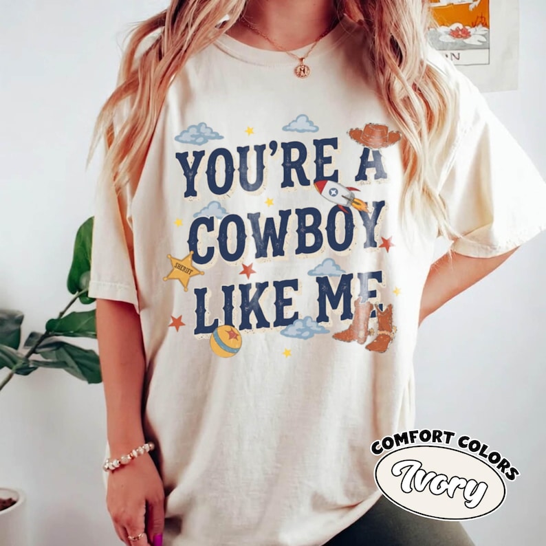 May include: An ivory short-sleeve t-shirt featuring the dark blue text "YOU'RE A COWBOY LIKE ME" surrounded by graphic illustrations of a rocket, a sheriff star, a ball, clouds, a cowboy hat, and a pair of brown cowboy boots in various colors.