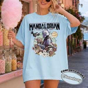 Op de afbeelding: Een lichtblauw t-shirt met een grafische print van The Mandalorian en Grogu. Het ontwerp bevat de tekst "The Mandalorian This Is The Way" boven de personages, omringd door een cirkelvormig kader, een planeet en diverse kleurrijke bloemen.
