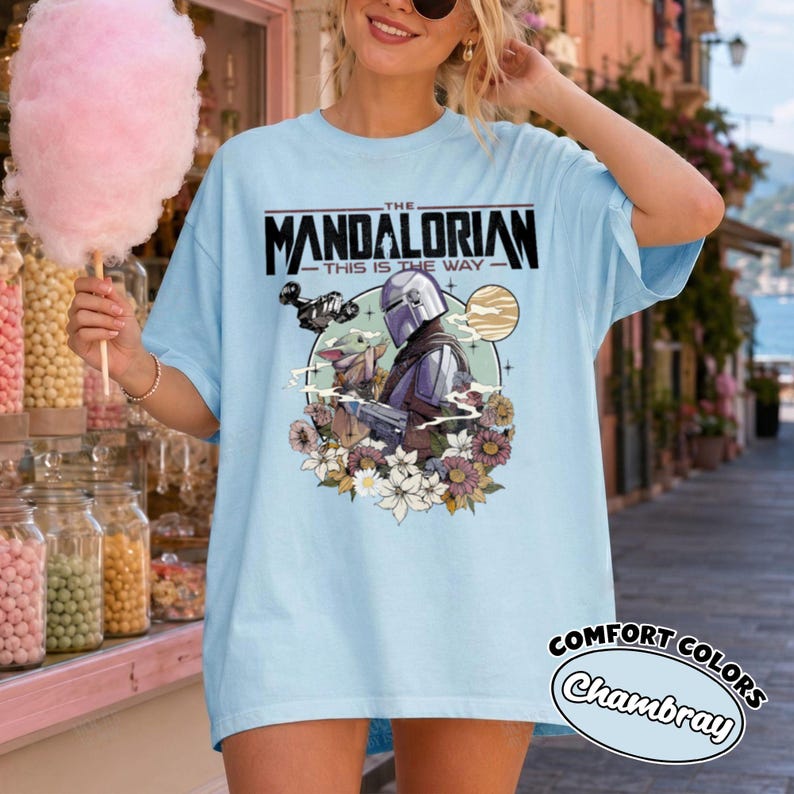 Op de afbeelding: Een lichtblauw t-shirt met een grafische print van The Mandalorian en Grogu. Het ontwerp bevat de tekst "The Mandalorian This Is The Way" boven de personages, omringd door een cirkelvormig kader, een planeet en diverse kleurrijke bloemen.