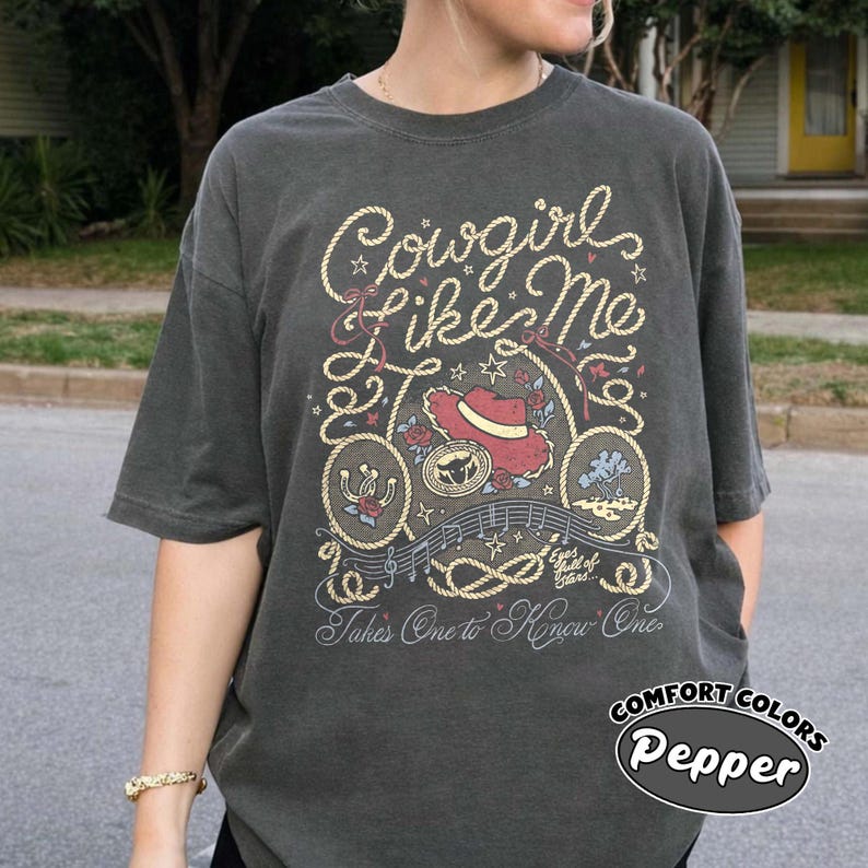 以下が含まれることがあります： チャコールグレーの半袖Tシャツ。ロープ風の文字で「Cowgirl Like Me」と「Takes One to Know One」と書かれたグラフィックデザインが特徴です。プリントには、赤いカウボーイハット、蹄鉄、バラ、音符がクリーム色で描かれています。