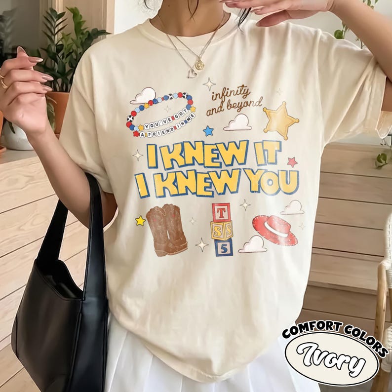 May include: An ivory short-sleeve t-shirt featuring a graphic print with the text "I KNEW IT I KNEW YOU", "infinity and beyond", and "YOU'VE GOT A FRIEND IN ME". The design includes a beaded bracelet, a star, cowboy boots, blocks, and a hat.