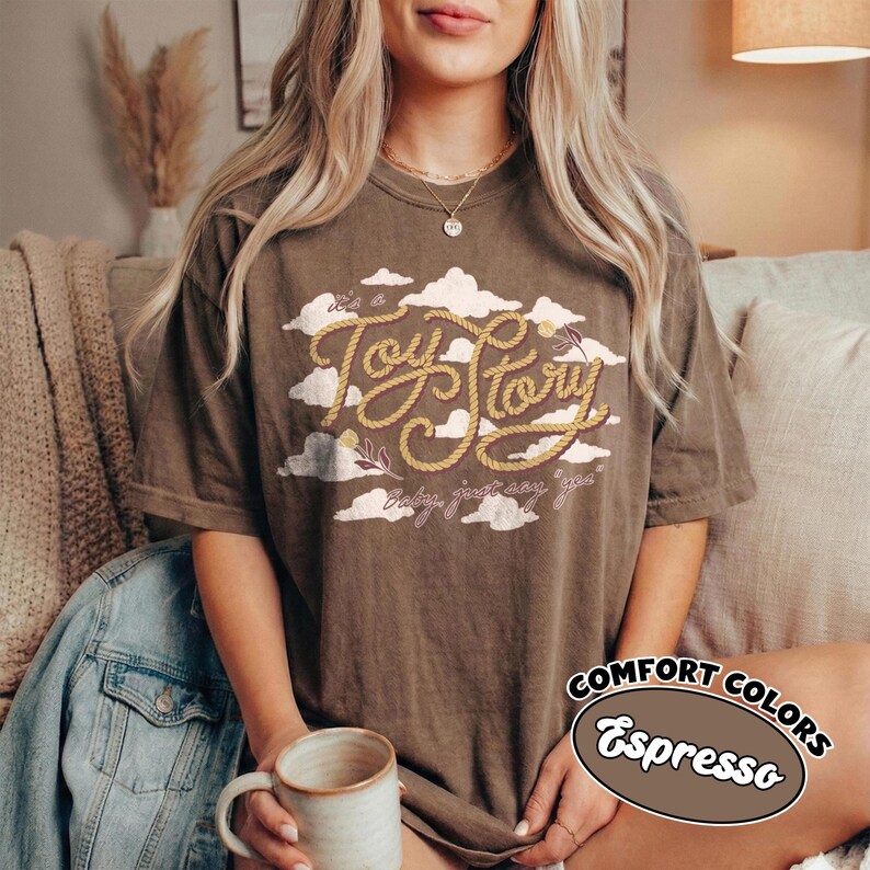 May include: An espresso-colored short-sleeve t-shirt featuring a graphic of white clouds and the text "It's a Toy Story" written in a rope-style font, with the additional text "baby, just say yes" printed below in a cursive script font.