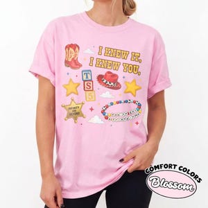 Peut inclure: Un t-shirt rose clair &agrave; manches courtes avec un imprim&eacute; graphique comprenant le texte I KNEW IT, I KNEW YOU, une botte de cow-boy rouge, un chapeau de cow-boy rouge, des &eacute;toiles jaunes, un badge de sh&eacute;rif, des cubes et un bracelet.