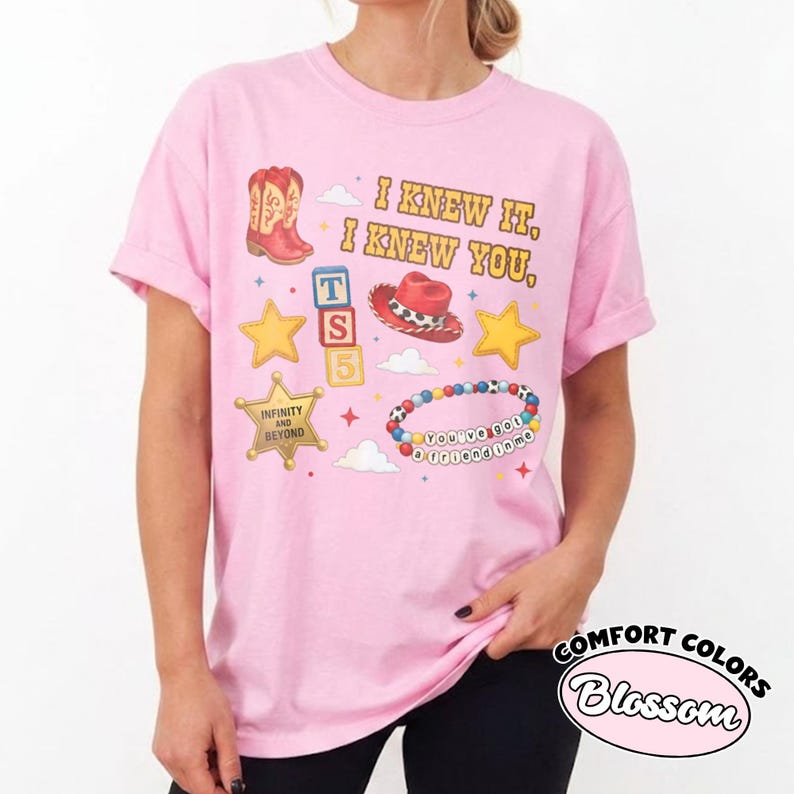 Peut inclure: Un t-shirt rose clair &agrave; manches courtes avec un imprim&eacute; graphique comprenant le texte I KNEW IT, I KNEW YOU, une botte de cow-boy rouge, un chapeau de cow-boy rouge, des &eacute;toiles jaunes, un badge de sh&eacute;rif, des cubes et un bracelet.