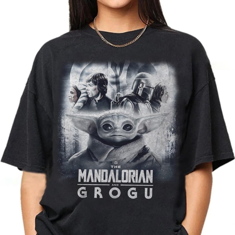 May include: Black t-shirt with a graphic of Grogu from The Mandalorian series. The design includes characters like Luke Skywalker and The Mandalorian. The text "THE MANDALORIAN and GROGU" is printed below the image.