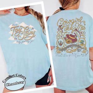 May include: A light blue t-shirt featuring a front graphic with the text "It's a Toy Story, baby, just say yes" in rope-style lettering over clouds. The back graphic displays "Cowgirl like me" and "Takes one to know one" with rope borders, a hat, and icons.