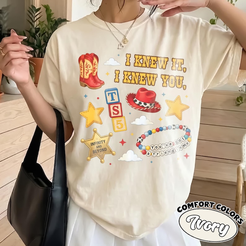 Peut inclure: Un t-shirt &agrave; manches courtes ivoire avec une impression graphique comprenant le texte I KNEW IT, I KNEW YOU, une botte de cowboy rouge, un chapeau de cowboy rouge, des blocs alphabet, des &eacute;toiles jaunes, un badge de sh&eacute;rif et un bracelet.