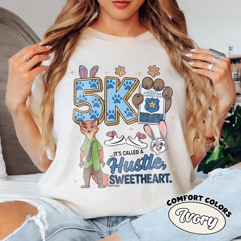 Puede incluir: Camiseta de color marfil con un dise&ntilde;o gr&aacute;fico que presenta el texto "5K" en azul con huellas de patas, personajes de dibujos animados y la frase "...It's called a Hustle, Sweetheart." La camiseta tambi&eacute;n tiene las palabras "Springtime Surprise 2024".