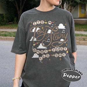 May include: A charcoal gray short-sleeve t-shirt featuring a graphic design with rope-style lettering that reads "ITS A Toy Story BABY JUST YES SAY". The print includes white clouds, small stars, musical notes, and colorful beads.