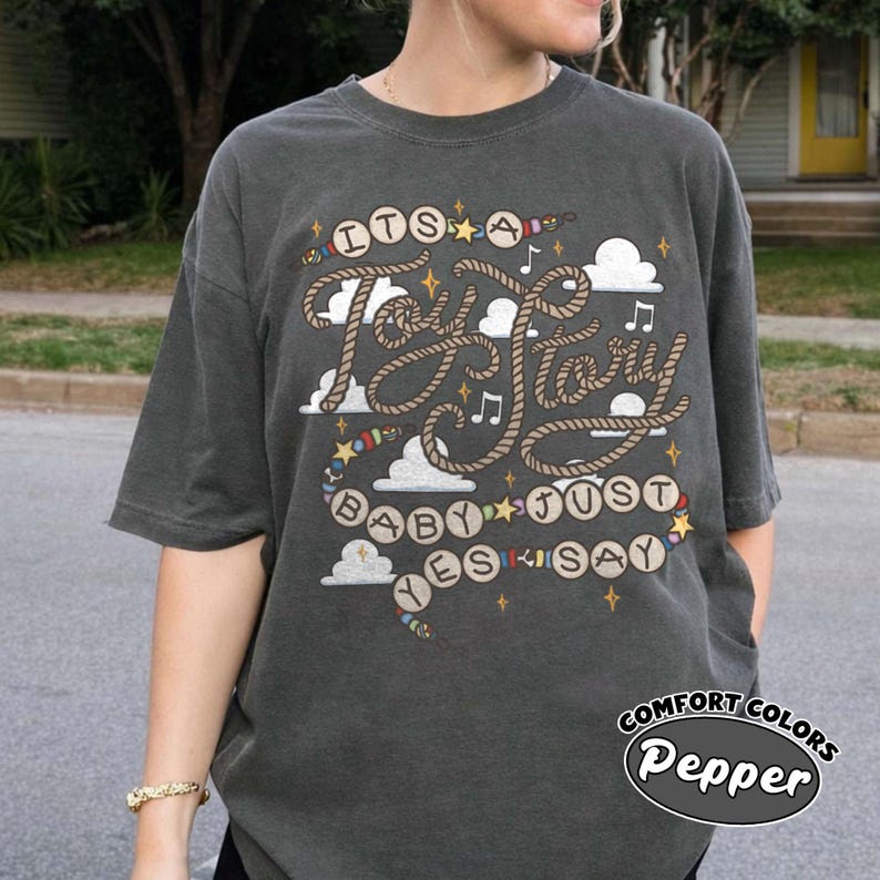 May include: A charcoal gray short-sleeve t-shirt featuring a graphic design with rope-style lettering that reads "ITS A Toy Story BABY JUST YES SAY". The print includes white clouds, small stars, musical notes, and colorful beads.