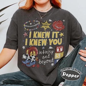 May include: A charcoal gray short-sleeve t-shirt featuring a graphic print with the text I KNEW IT I KNEW YOU and infinity and beyond. The design includes a sheriff badge, a red cowboy hat, cowboy boots, and alphabet blocks with letters T and S.