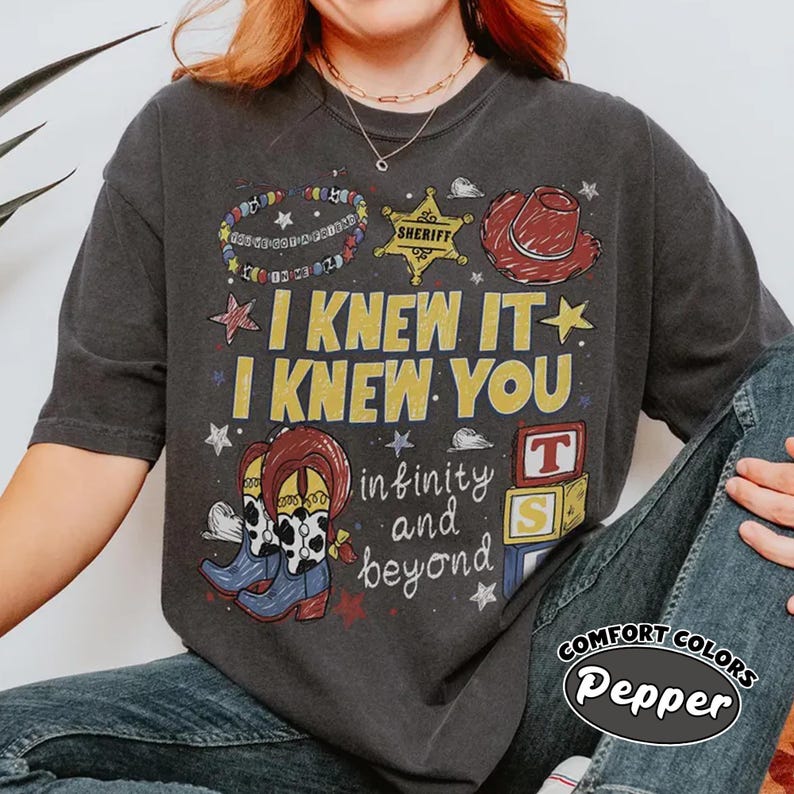 May include: A charcoal gray short-sleeve t-shirt featuring a graphic print with the text I KNEW IT I KNEW YOU and infinity and beyond. The design includes a sheriff badge, a red cowboy hat, cowboy boots, and alphabet blocks with letters T and S.