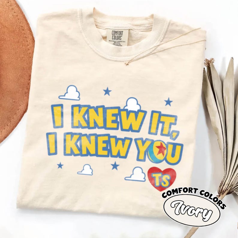 May include: An ivory short-sleeve t-shirt with a crew neckline. The graphic features the text "I KNEW IT, I KNEW YOU" in yellow with blue outlines, accompanied by blue stars, white cloud shapes, a colorful ball icon, and a red heart with "TS" inside.