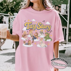 May include: A light pink t-shirt featuring the text Tay Story in yellow with a blue outline. The graphic includes multiple illustrated figures dressed in various outfits, each sitting on a white cloud, surrounded by small yellow stars and musical notes.