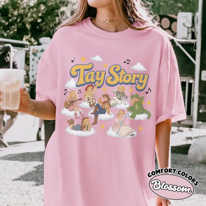 May include: A light pink t-shirt featuring the text Tay Story in yellow with a blue outline. The graphic includes multiple illustrated figures dressed in various outfits, each sitting on a white cloud, surrounded by small yellow stars and musical notes.