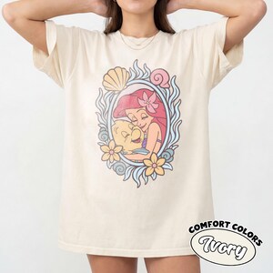 May include: An ivory short-sleeve t-shirt featuring a central graphic print of a red-haired character embracing a yellow fish. The illustration is framed by blue swirling patterns, yellow flowers, and seashells. The text Comfort Colors Ivory is visible.