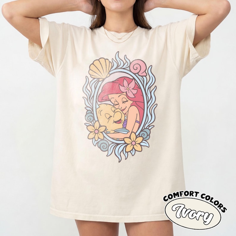 May include: An ivory short-sleeve t-shirt featuring a central graphic print of a red-haired character embracing a yellow fish. The illustration is framed by blue swirling patterns, yellow flowers, and seashells. The text Comfort Colors Ivory is visible.