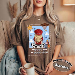May include: A brown t-shirt featuring a graphic of a blonde character wearing a red cowboy hat and shirt, sitting with cow-print pants. The design includes a brown cowboy hat, stars, alphabet blocks, and the text I KNEW IT I KNEW YOU.