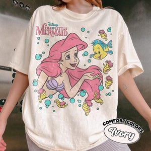 Puede incluir: Una camiseta de manga corta color marfil con un gr&aacute;fico de Ariel de La Sirenita con cabello rosa, un top de conchas morado y Flounder. El dise&ntilde;o incluye caballitos de mar amarillos y burbujas turquesas, con el texto Disney The Little Mermaid.