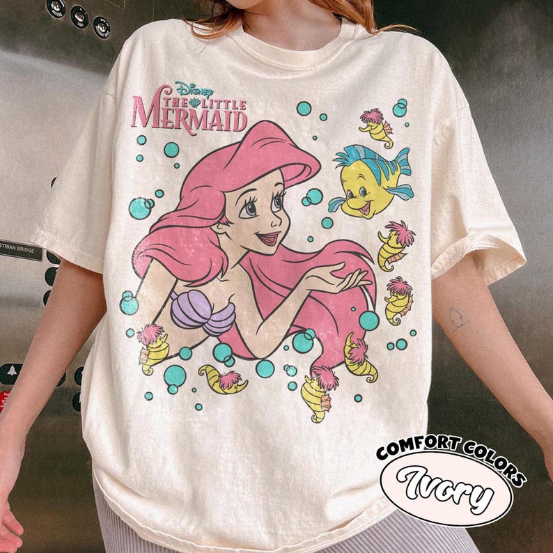 Puede incluir: Una camiseta de manga corta color marfil con un gr&aacute;fico de Ariel de La Sirenita con cabello rosa, un top de conchas morado y Flounder. El dise&ntilde;o incluye caballitos de mar amarillos y burbujas turquesas, con el texto Disney The Little Mermaid.