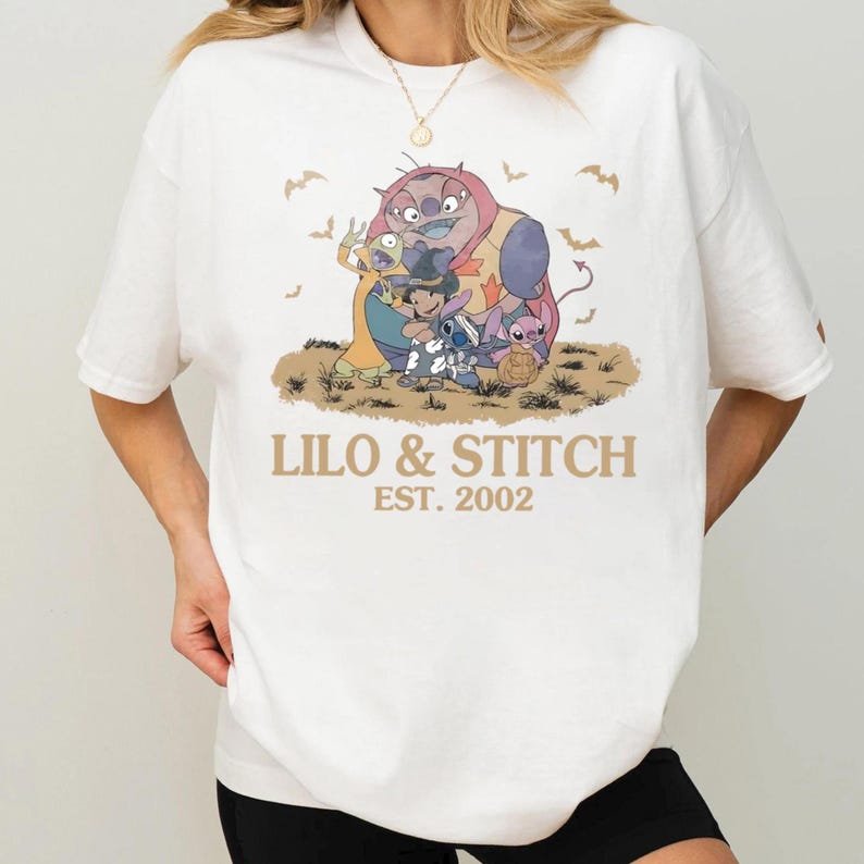 K&ouml;nnte beinhalten: Wei&szlig;es T-Shirt mit einem goldenen Aufdruck von Lilo & Stitch-Charakteren und dem Text "Lilo & Stitch Est. 2002". Das Design umfasst Cartoonfiguren und Flederm&auml;use. Ein l&auml;ssiges Kleidungsst&uuml;ck.