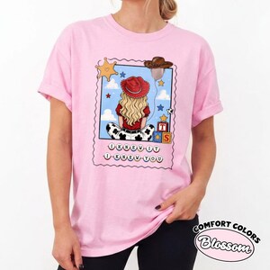 May include: A light pink t-shirt featuring a graphic print of a blonde person wearing a red cowboy hat, sitting with their back to the viewer. The design includes a sheriff star, a brown hat, toy blocks, and the text I KNEW IT I KNEW YOU.