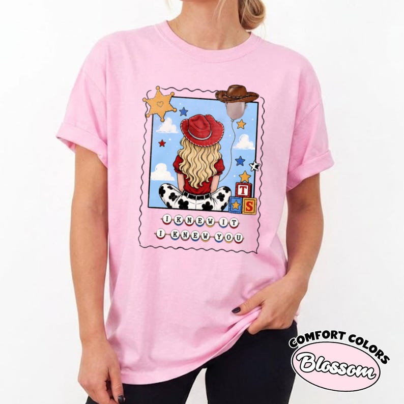 May include: A light pink t-shirt featuring a graphic print of a blonde person wearing a red cowboy hat, sitting with their back to the viewer. The design includes a sheriff star, a brown hat, toy blocks, and the text I KNEW IT I KNEW YOU.