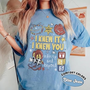 May include: A blue t-shirt featuring a graphic print with the text I KNEW IT I KNEW YOU and infinity and beyond. The design includes a sheriff badge, a cowboy hat, a beaded bracelet, cowboy boots, and three stacked blocks labeled T, S, and 5.