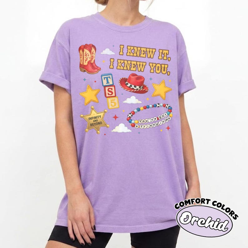 Peut inclure: Un t-shirt violet clair avec une impression graphique comprenant le texte I KNEW IT, I KNEW YOU, une botte de cowboy rouge, un chapeau de cowboy rouge, des &eacute;toiles jaunes, des blocs avec T, S, 5, un badge de sh&eacute;rif et un bracelet de perles.
