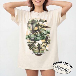 Pu&ograve; includere: T-shirt color avorio con grafica di "The Mandalorian" con il testo "The Mandalorian and Grogu" e "This is the Way". Il design include immagini del Mandaloriano e Grogu. La maglietta &egrave; etichettata "Comfort Colors Ivory".