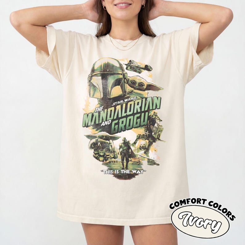 Pu&ograve; includere: T-shirt color avorio con grafica di "The Mandalorian" con il testo "The Mandalorian and Grogu" e "This is the Way". Il design include immagini del Mandaloriano e Grogu. La maglietta &egrave; etichettata "Comfort Colors Ivory".