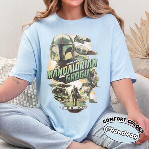 Pu&ograve; includere: T-shirt azzurra con grafica della serie Star Wars, "The Mandalorian and Grogu." Il design include immagini del Mandaloriano, Grogu e un'astronave, con la frase "This is the Way."