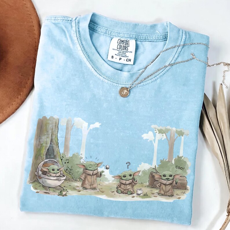 Puede incluir: Camiseta Comfort Colors azul claro con una escena de dibujos animados de Grogu. El dise&ntilde;o muestra a Grogu en una c&aacute;psula, con una nota musical, una pelota y un signo de interrogaci&oacute;n. Tambi&eacute;n se ve un collar plateado con un colgante.