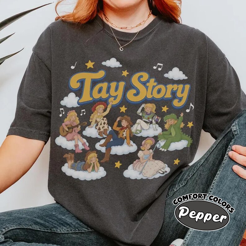 May include: A charcoal gray short-sleeve t-shirt featuring the text Tay Story in yellow with a blue outline. The graphic includes seven illustrated figures dressed as characters on white clouds, surrounded by yellow stars and musical notes.