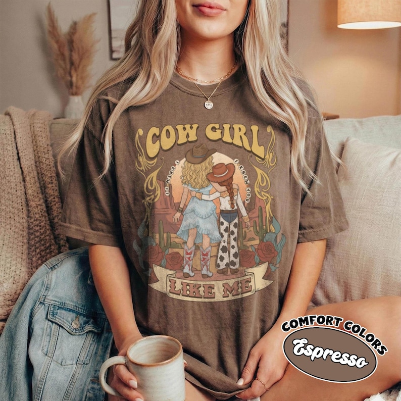 May include: A brown short-sleeve t-shirt featuring a graphic print of two figures in cowboy hats standing in a desert landscape with cacti and roses. The text reads COW GIRL LIKE ME. The shirt has a vintage-style wash and a crew neckline.