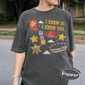 Peut inclure: Un t-shirt gris anthracite &agrave; manches courtes avec un imprim&eacute; graphique repr&eacute;sentant une botte de cowboy rouge, un chapeau de cowboy rouge, des &eacute;toiles jaunes, des blocs alphab&eacute;tiques et un bracelet de perles color&eacute;. Le texte indique I KNEW IT, I KNEW YOU, INFINITY AND BEYOND, et YOU'VE GOT A FRIEND IN ME.