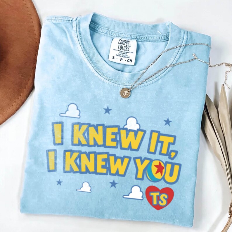 May include: A light blue crew neck t-shirt featuring the yellow text "I KNEW IT, I KNEW YOU" with a small colorful ball graphic, a red heart containing the yellow letters "TS", and white cloud and blue star graphics printed on the front.