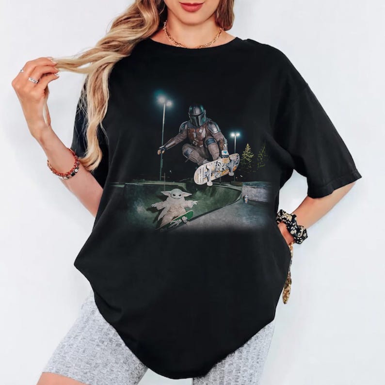 May include: Black t-shirt featuring a graphic of The Mandalorian and Grogu skateboarding in a skate park at night. The design includes a detailed illustration of the characters and skateboards, set against a backdrop of streetlights and trees.