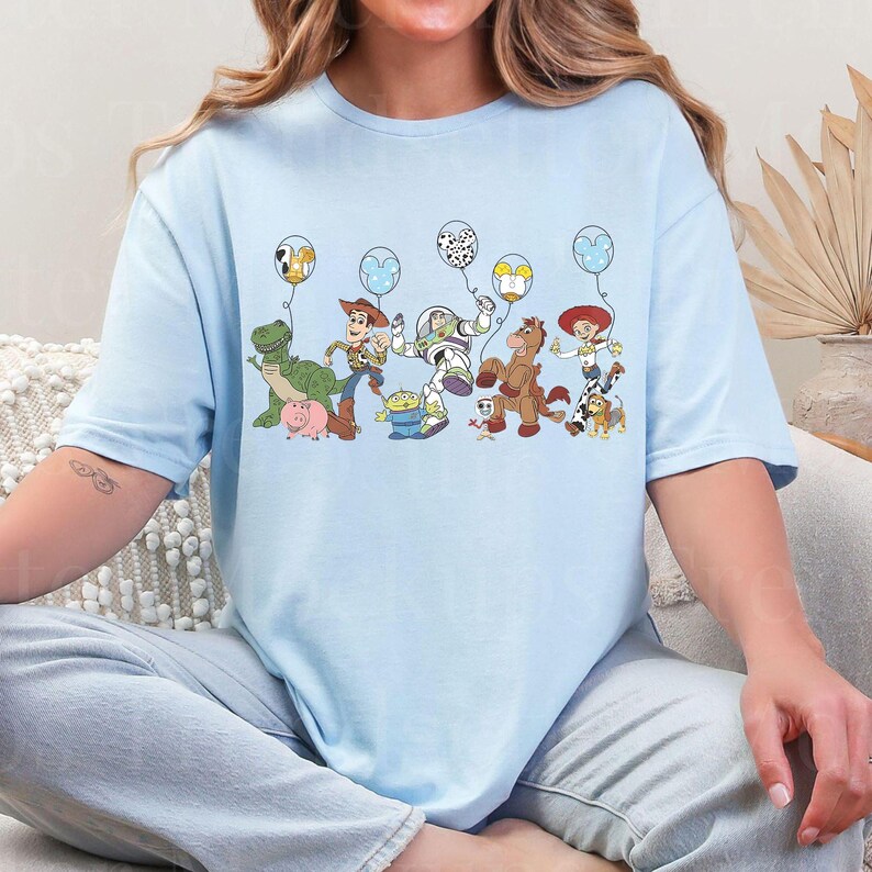 May include: Pale blue t-shirt with a vibrant graphic of animated characters holding balloons. The design includes a dinosaur, a pig, a cowboy, an astronaut, a horse, and other characters. The shirt is made of a soft material.