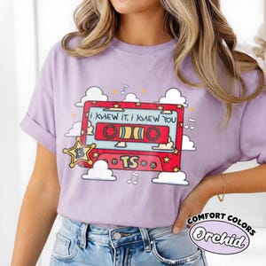 May include: A light purple t-shirt featuring a graphic of a red cassette tape with the text "I KNEW IT, I KNEW YOU" and "TS". The design includes a yellow star with the number 13, white clouds, and musical notes surrounding the cassette tape.