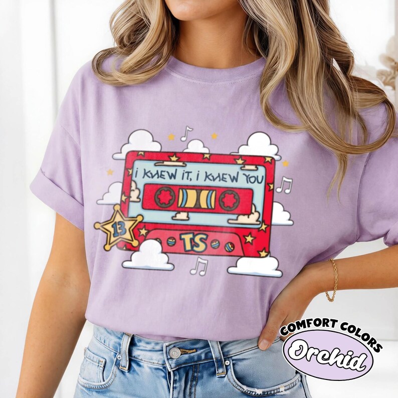 May include: A light purple t-shirt featuring a graphic of a red cassette tape with the text "I KNEW IT, I KNEW YOU" and "TS". The design includes a yellow star with the number 13, white clouds, and musical notes surrounding the cassette tape.