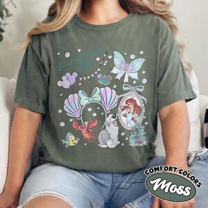 May include: A moss green short-sleeve t-shirt featuring a graphic print of a mermaid, a crab, a fish, a dog, a butterfly, and a pair of seashell ears. The text Part of Your World is printed in teal script above the illustrations with small bubbles.