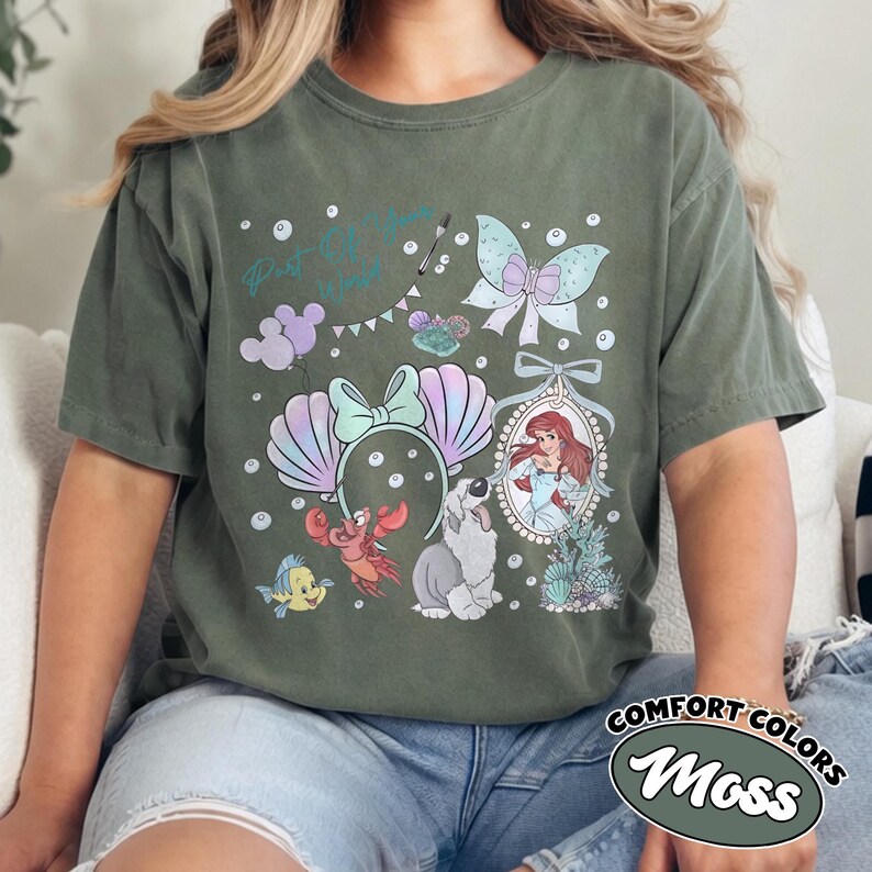 May include: A moss green short-sleeve t-shirt featuring a graphic print of a mermaid, a crab, a fish, a dog, a butterfly, and a pair of seashell ears. The text Part of Your World is printed in teal script above the illustrations with small bubbles.