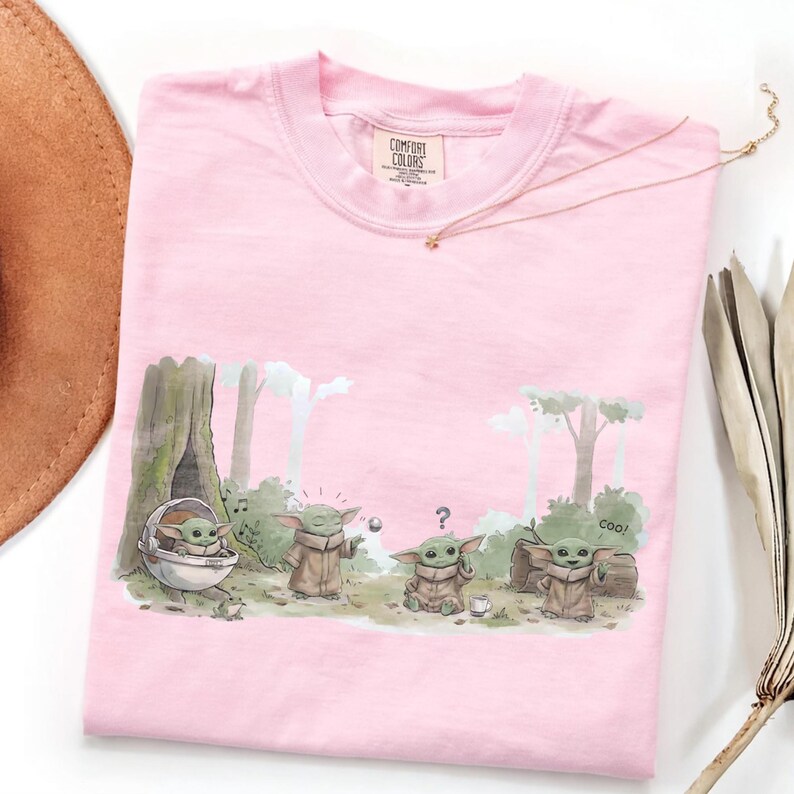 Puede incluir: Camiseta Comfort Colors rosa p&aacute;lido con una ilustraci&oacute;n de dibujos animados de un personaje de una popular serie de ciencia ficci&oacute;n. El gr&aacute;fico muestra al personaje en un entorno forestal. Un collar dorado est&aacute; drapeado sobre la camiseta.
