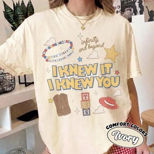 May include: An ivory t-shirt featuring a graphic print with the text "I KNEW IT I KNEW YOU", "infinity and beyond", and "YOU'VE GOT A FRIEND IN ME". The design includes a beaded bracelet, a sheriff star, cowboy boots, alphabet blocks, and a hat.