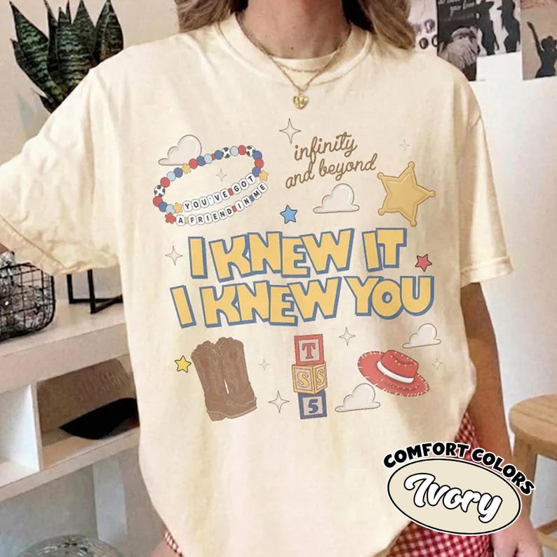 May include: An ivory t-shirt featuring a graphic print with the text "I KNEW IT I KNEW YOU", "infinity and beyond", and "YOU'VE GOT A FRIEND IN ME". The design includes a beaded bracelet, a sheriff star, cowboy boots, alphabet blocks, and a hat.